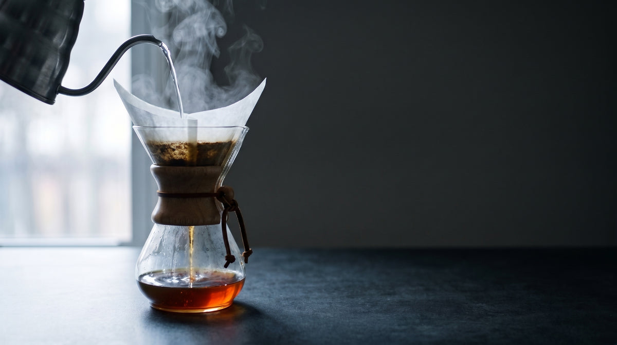 Hot water poured from a gooseneck kettle into a Chemex of coffee, with steam rising